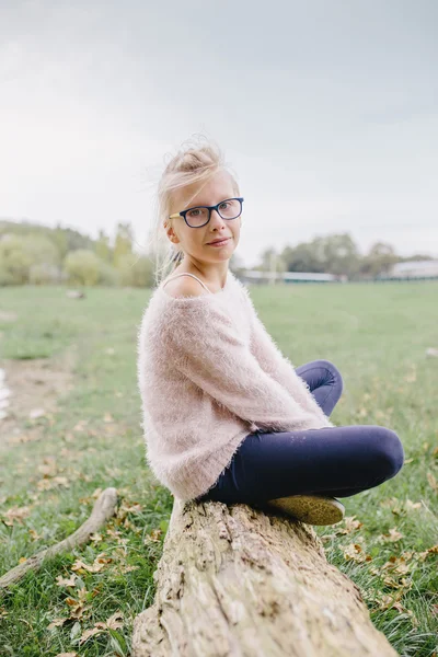 Blond tjej med glasögon sittande med korsade ben på en stock utomhus — Stockfoto