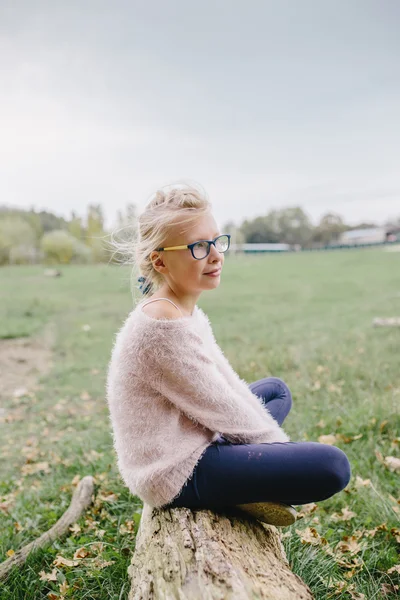 Blondes Mädchen mit Brille sitzt mit überkreuzten Beinen auf einem Baumstamm im Freien — Stockfoto