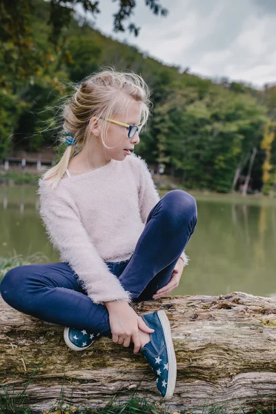 Blond tjej med glasögon sitter med korsade ben på en stock utomhus på bakgrunden av sjön — Stockfoto
