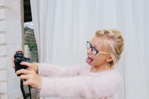 Blond tjej med glasögon som visar tungan och fotograferar sig själv på kameran. Flickan gör Selfie på kameran på bakgrunden av vita gardin — Stockfoto