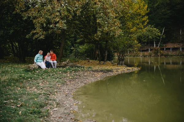 Chica, hombre joven en un suéter rojo y una anciana sentada en un tronco junto al lago. hija, padre y abuela se divierten al aire libre en el parque — Foto de Stock