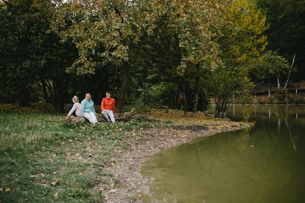 Chica, hombre joven en un suéter rojo y una anciana sentada en un tronco junto al lago. hija, padre y abuela se divierten al aire libre en el parque — Foto de Stock