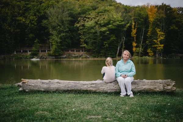 A menina virou as costas à mulher mais velha. menina ofendida e uma mulher idosa sentado em um log no fundo do lago. conflito de gerações. problemas familiares . — Fotografia de Stock