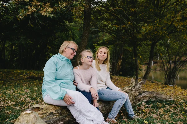 Menina, mulher jovem e mulher madura sentado em um log no parque. Filha, mãe e avó relaxar ao ar livre no fundo da floresta de outono e lago — Fotografia de Stock