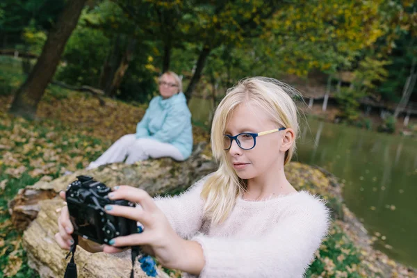 Chica rubia con gafas se fotografió en la cámara al aire libre en el fondo del lago y su abuela. Chica hace selfie en cámara en el parque — Foto de Stock