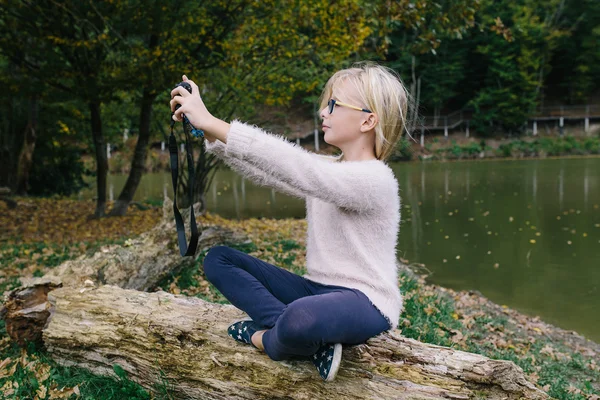 Blond tjej med glasögon fotograferade sig själv på kamera utomhus på bakgrunden av sjön. Flicka gör selfie på kameran i park — Stockfoto