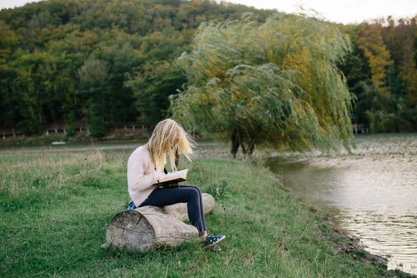 Pequena loira desarrumada sentada em um tronco junto ao lago e lendo um livro — Fotografia de Stock