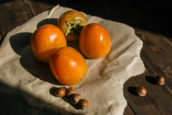 ripe persimmon lies on kraft paper on brown wooden background, near lies group of brown hazel nut. Vegetarian nutrition. Healthy eating, dieting