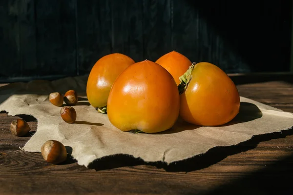 Ripe persimmon lies on kraft paper on brown wooden background, near lies group of brown hazel nut. Vegetarian nutrition. Healthy eating, dieting — Stock Photo, Image