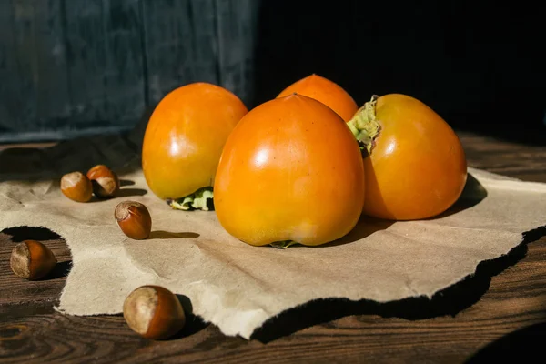 Ripe persimmon lies on kraft paper on brown wooden background, near lies group of brown hazel nut. Vegetarian nutrition. Healthy eating, dieting — Stock Photo, Image