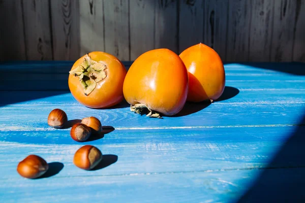 Ripe persimmon on a blue wooden background, near lies group of brown hazel nut. Vegetarian nutrition.  Healthy eating, dietin — Stock Photo, Image