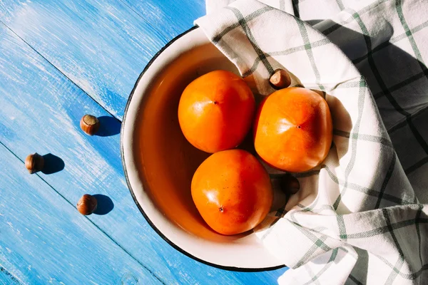 Ripe persimmon is in a bowl with a cloth napkins on a blue background, near lies group of brown hazel nut. Vegetarian nutrition. — Stock Photo, Image