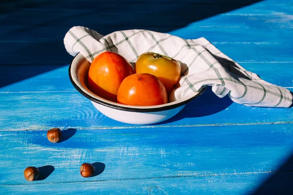 Ripe persimmon is in a bowl with a cloth napkins on a blue background, near lies group of brown hazel nut. Vegetarian nutrition. Vegetarian nutrition. Healthy eating, dieting — Stock Photo, Image