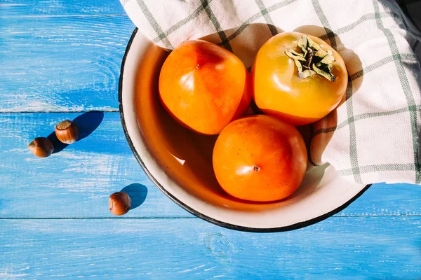 Ripe persimmon is in a bowl with a cloth napkins on a blue background, near lies group of brown hazel nut. Vegetarian nutrition. Vegetarian nutrition. Healthy eating, dieting — Stock Photo, Image