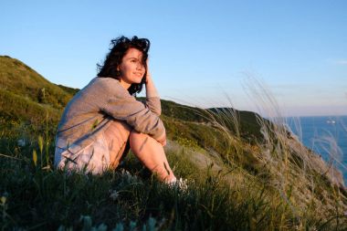 tepe üzerinde çimenlerin üzerinde deniz üzerine oturan güzel esmer kadın. Kadın Okyanusu yakınlarındaki sırt çantası ile seyahat. Seyahat; yaşam tarzı kavramı 