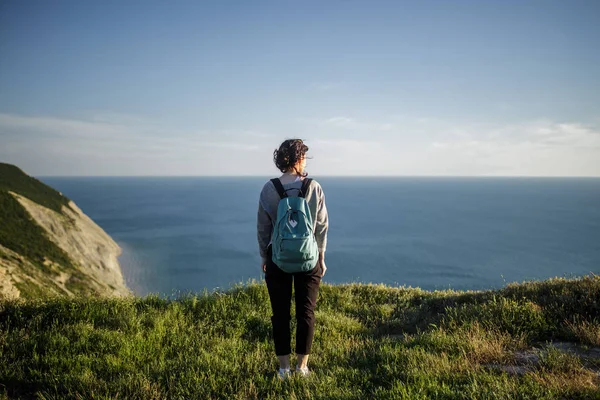 年轻女子徒步旅行者带着背包站在悬崖上 期待着大海 天空的背景 游客在山顶上欣赏风景 自由和积极的生活方式概念 — 图库照片