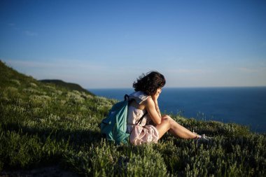 tepe üzerinde çimenlerin üzerinde deniz üzerine oturan güzel esmer kadın. Kadın Okyanusu yakınlarındaki sırt çantası ile seyahat. Seyahat, yaşam kavramı