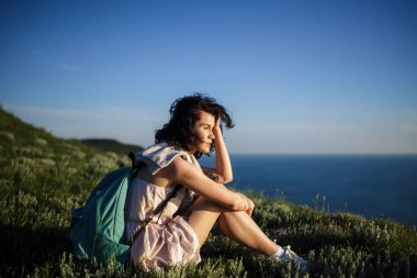 tepe üzerinde çimenlerin üzerinde deniz üzerine oturan güzel esmer kadın. Kadın Okyanusu yakınlarındaki sırt çantası ile seyahat. Seyahat, yaşam kavramı