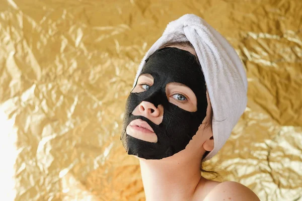 Jonge Vrouw Toe Passen Zwarte Cosmetische Stof Gezichtsmasker Gouden Achtergrond — Stockfoto