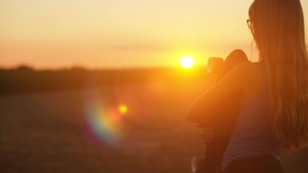 Chica sosteniendo monopatín y mirando la puesta de sol — Vídeo de stock