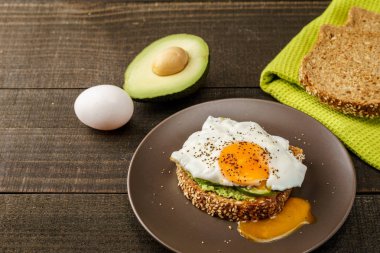 avokado ve yeşil mutfak havlu bir rustik ahşap ve tablo ekmek üzerinde kahverengi bir tabak kızarmış yumurta sandviç.
