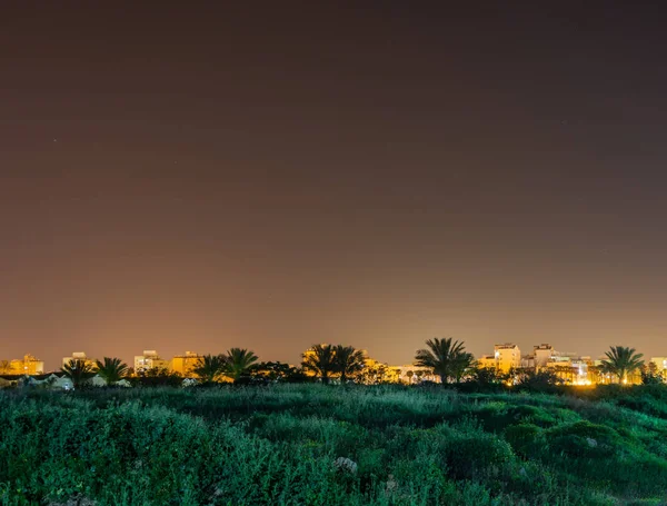 El campo sin tratar con la hierba verde jugosa con la ciudad en el fondo en Israel por la noche . — Foto de Stock