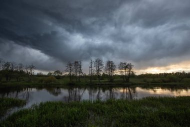 Nehir manzarası, ön planda yeşil çimenleri görebilirsiniz. Sonra kıyıda duran ağaçları görebilirsiniz, ve gökyüzünde fırtına bulutları vardır, sağ köşede küçük güneş boşlukları vardır.