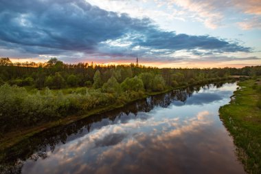 Bu fotoğrafta, çerçevenin büyük bir kısmı fırtınalı gökyüzü tarafından nehrin sakin suyuna yansıtılır. Sağda pembe bulutlar ve solda koyu mavi bulutlar. Sol tarafta yoğun bir orman yetişiyor. Gün batımı..