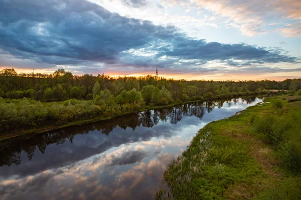 Bu fotoğrafta, çerçevenin büyük bir kısmı fırtınalı gökyüzü tarafından nehrin sakin suyuna yansıtılır. Sağda pembe bulutlar ve solda koyu mavi bulutlar. Sol tarafta yoğun bir orman yetişiyor. Gün batımı..
