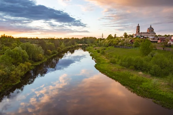 Çerçevenin altında bulutların ve açık mavi gökyüzünün yansıdığı sularda geniş ve sakin bir nehir görebilirsiniz. Sol tarafta sık bir orman yetişiyor ve sağ tarafta ağaçlar ve bir köy görülüyor. Tepede küçük bir şey görebilirsiniz. 