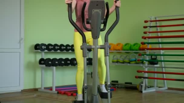 Jeune femme en forme utilisant un entraîneur elliptique dans un centre de remise en forme, écoutant une musique avec écouteurs et souriant. Portrait de fille de fitness dans la salle de gym près d'une fenêtre,. 4k — Video