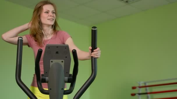 Ung passar kvinna med en elliptisk tränare i ett fitnesscenter, en musik med hörlurar och leende. Porträtt av fitness flicka i gymmet nära ett fönster. 4k — Stockvideo
