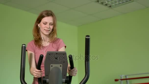 Mujer joven en forma usando un entrenador elíptico en un gimnasio, escuchando una música con auriculares y sonriendo. Retrato de chica fitness en el gimnasio cerca de una ventana,. 4k — Vídeos de Stock