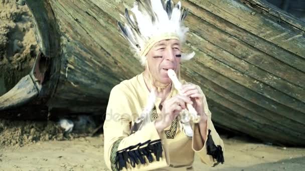 Ein Indianer in Nationaltracht spielt eine Flöte in der Nähe eines Bootes am Flussufer 4k — Stockvideo