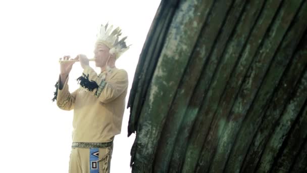 Un indio con un traje nacional toca una flauta cerca de un barco en la orilla del río 4k — Vídeo de stock