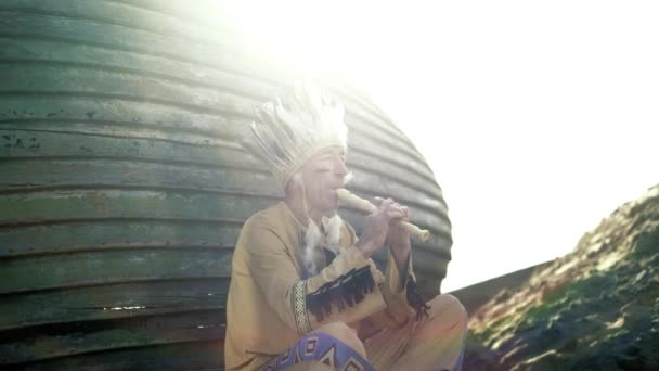 Un indio con un traje nacional toca una flauta cerca de un barco en la orilla del río 4k — Vídeos de Stock