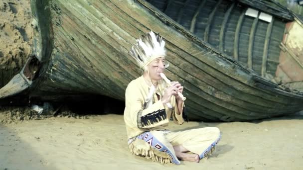 Un indio con un traje nacional toca una flauta cerca de un barco en la orilla del río 4k — Vídeos de Stock