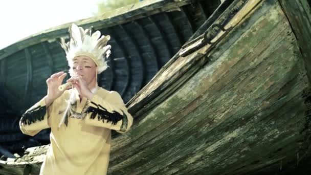 Indián v národním kroji hraje na flétnu v blízkosti lodi na břehu řeky 4k — Stock video