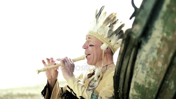 Indián v národním kroji hraje na flétnu v blízkosti lodi na břehu řeky 4k — Stock video