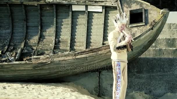 Un indiano in costume nazionale suona un flauto vicino a una barca sulla riva del fiume 4k — Video Stock