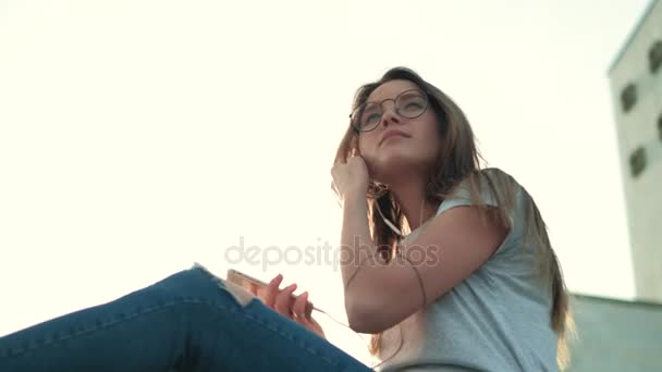 Siedzi brunetka dziewczyna w okularach o zachodzie słońca słucha muzyki — Wideo stockowe
