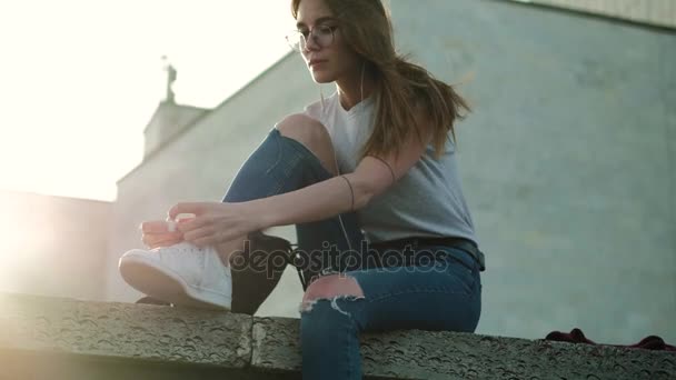 Menina morena em óculos senta-se ao pôr do sol escuta música amarra atacadores — Vídeo de Stock
