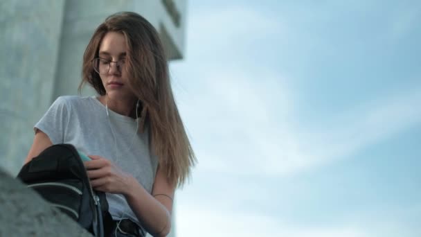 Brunett flicka i glasögon sitter vid solnedgången lyssnar på musik som letar efter något i en ryggsäck närbild — Stockvideo