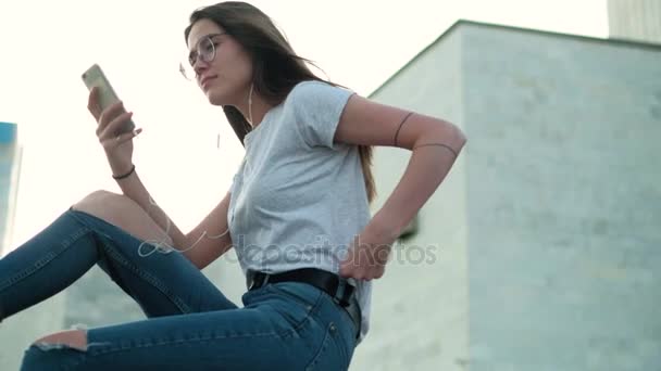 Brunette girl in glasses sits at sunset listens to music and reads messages — Stock Video