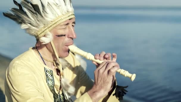 Un indio con un traje nacional toca una flauta en la orilla del río 4k — Vídeo de stock