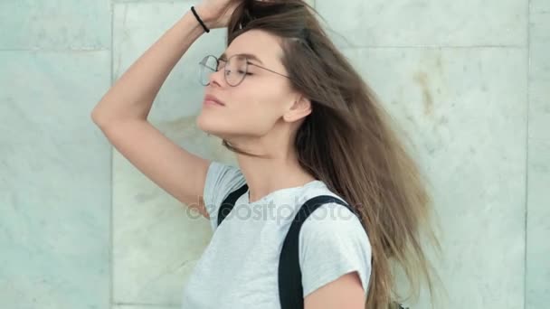 아름 다운 패션 여자 도시의 회색 코트에 거리에서 벽돌 벽 근처에 서. 갈색 섹시 한 여자 긴 머리와 안경 벽 근처 카메라 시계. 4 k — 비디오