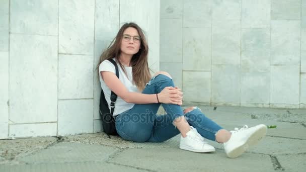 Flicka brunett i glas med en svart ryggsäck sitter på en grå marmor vägg, rätar ut håret och skrattar, jublar — Stockvideo