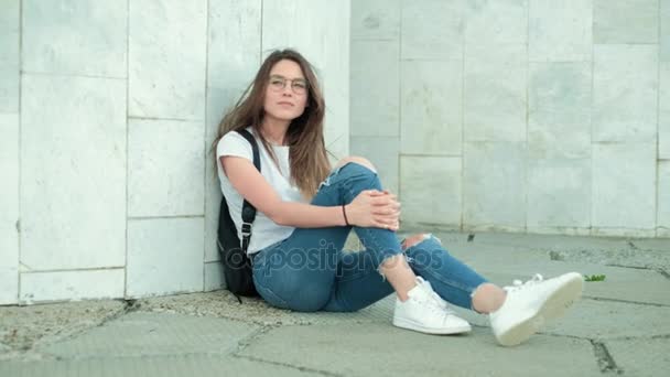 Chica morena en gafas con una mochila negra se sienta en una pared de mármol gris, endereza su cabello y se ríe, se regocija — Vídeo de stock