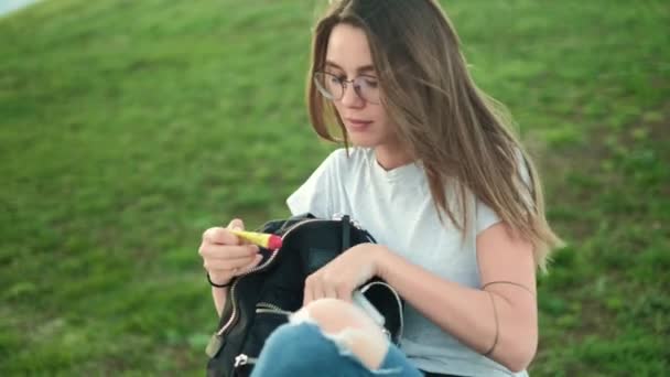 Menina morena bonita com óculos senta-se no parque na grama puxa para fora da mochila batom e hidrata-los — Vídeo de Stock