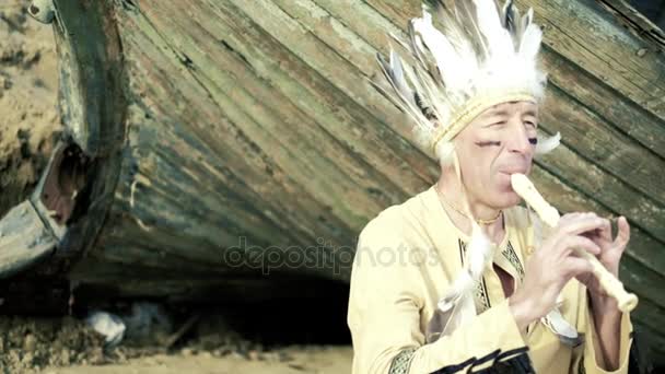 En Indian i en folkdräkt spelar en flöjt nära en båt på flodbank 4k — Stockvideo